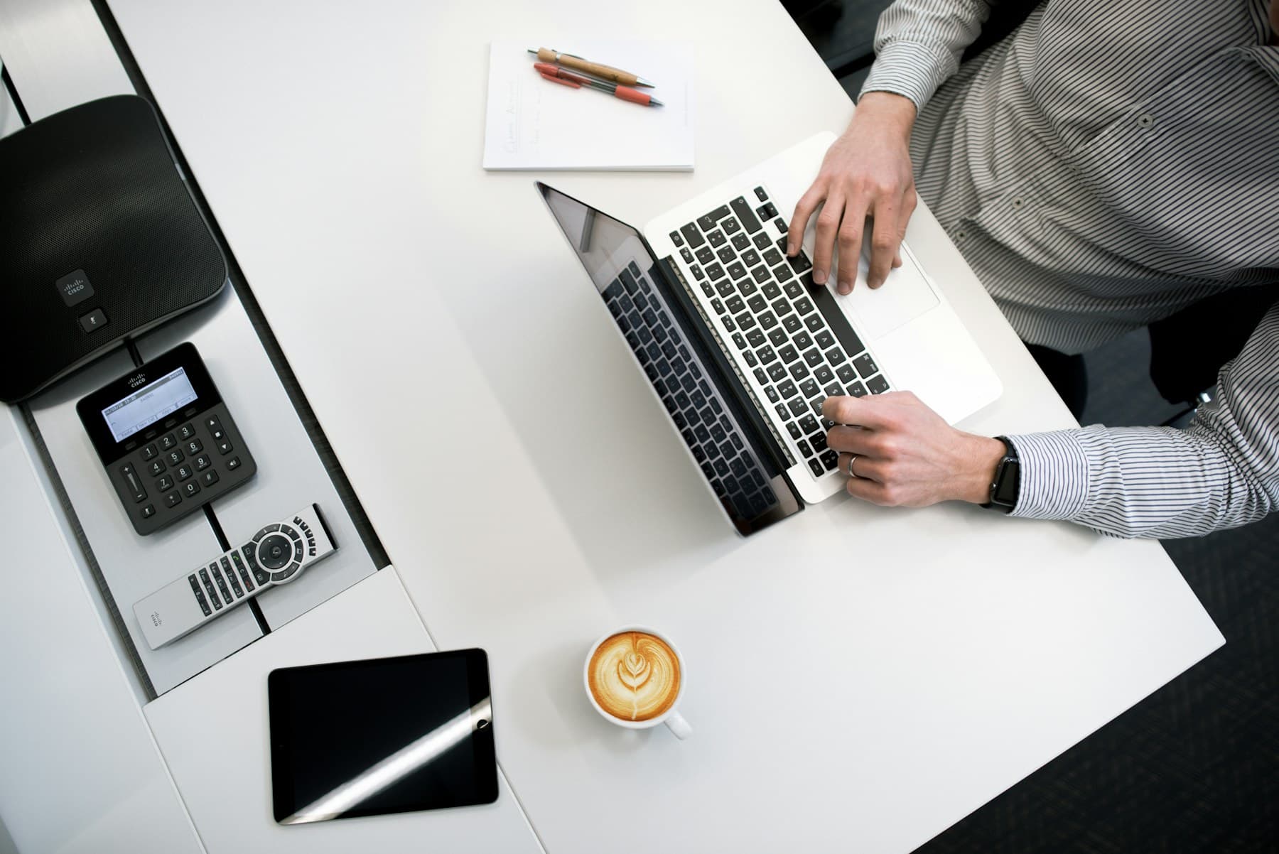 Overhead view of a white work desk with a laptop, a conference phone, a tablet, pens on a notepad and a cup of coffee