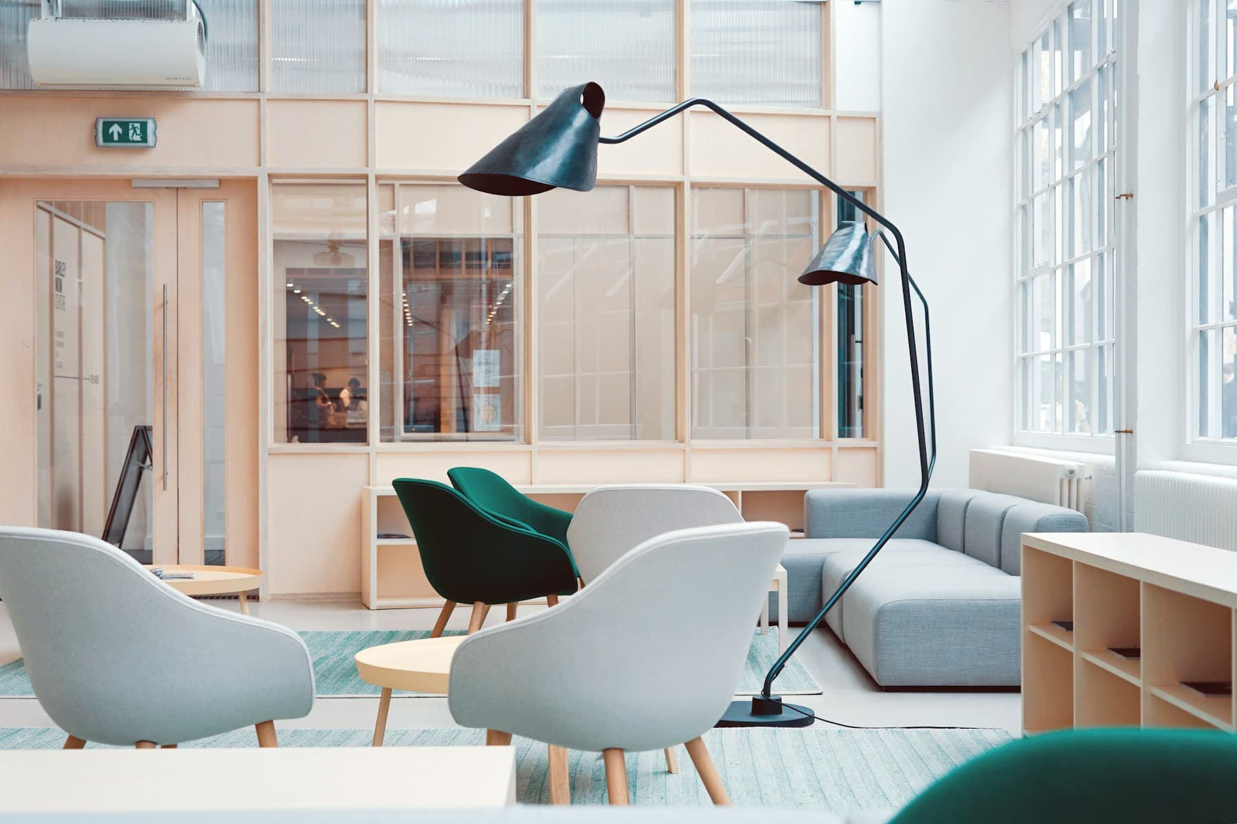 A bright office lounge with pale armchairs, a grey modular sofa and a tall two-headed floor lamp beside a glazed partition wall