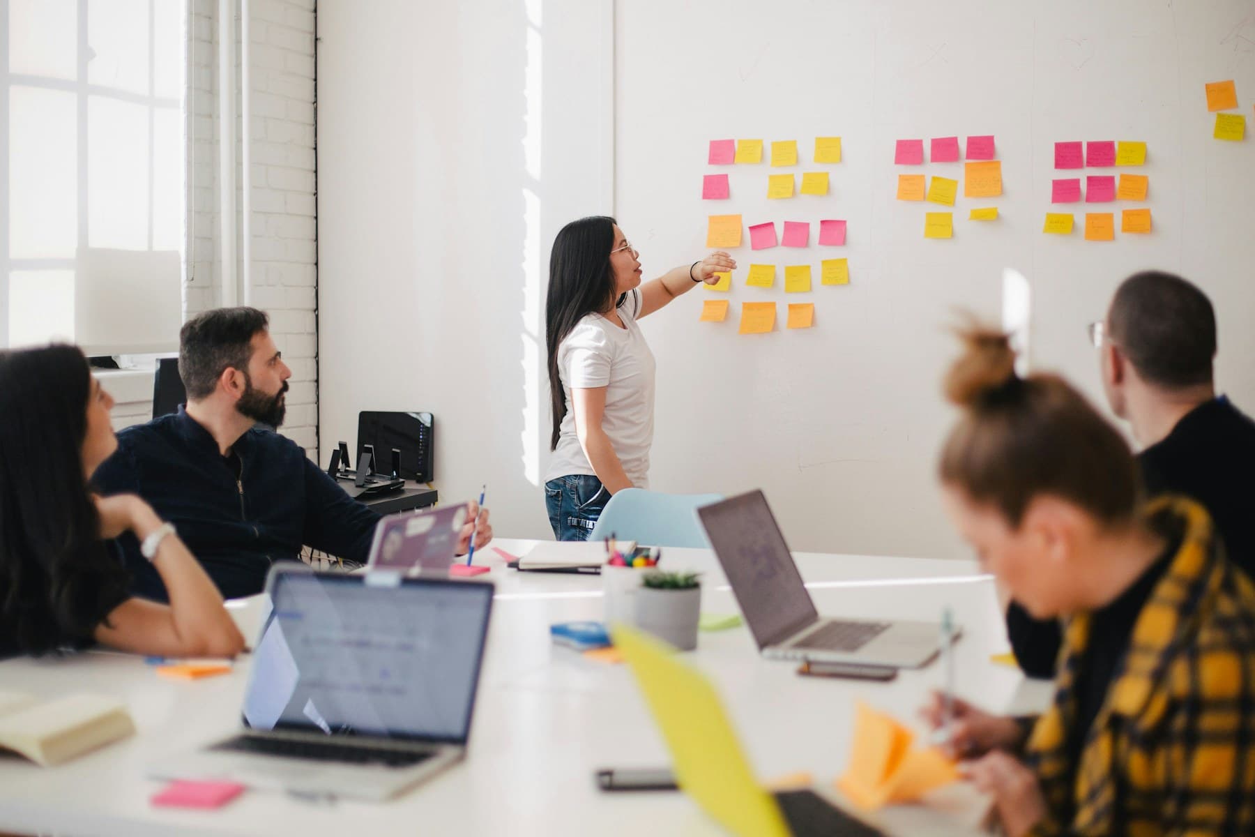 A woman pressing a sticky note onto a wall grid of pink, orange and yellow notes while colleagues watch from a table of open laptops