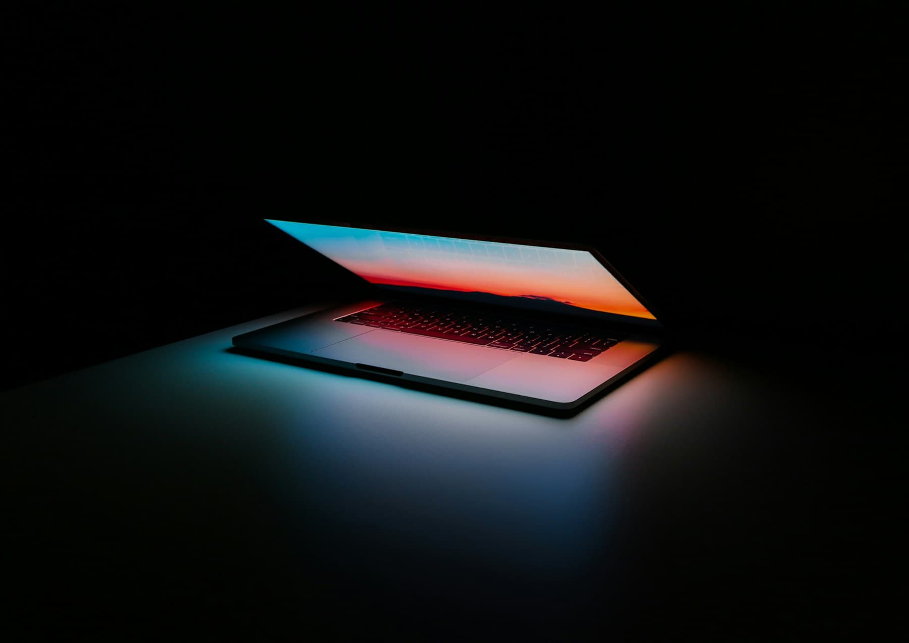 A half-open laptop in a dark room, the orange and blue gradient of its screen spilling across the desk and lighting the keyboard
