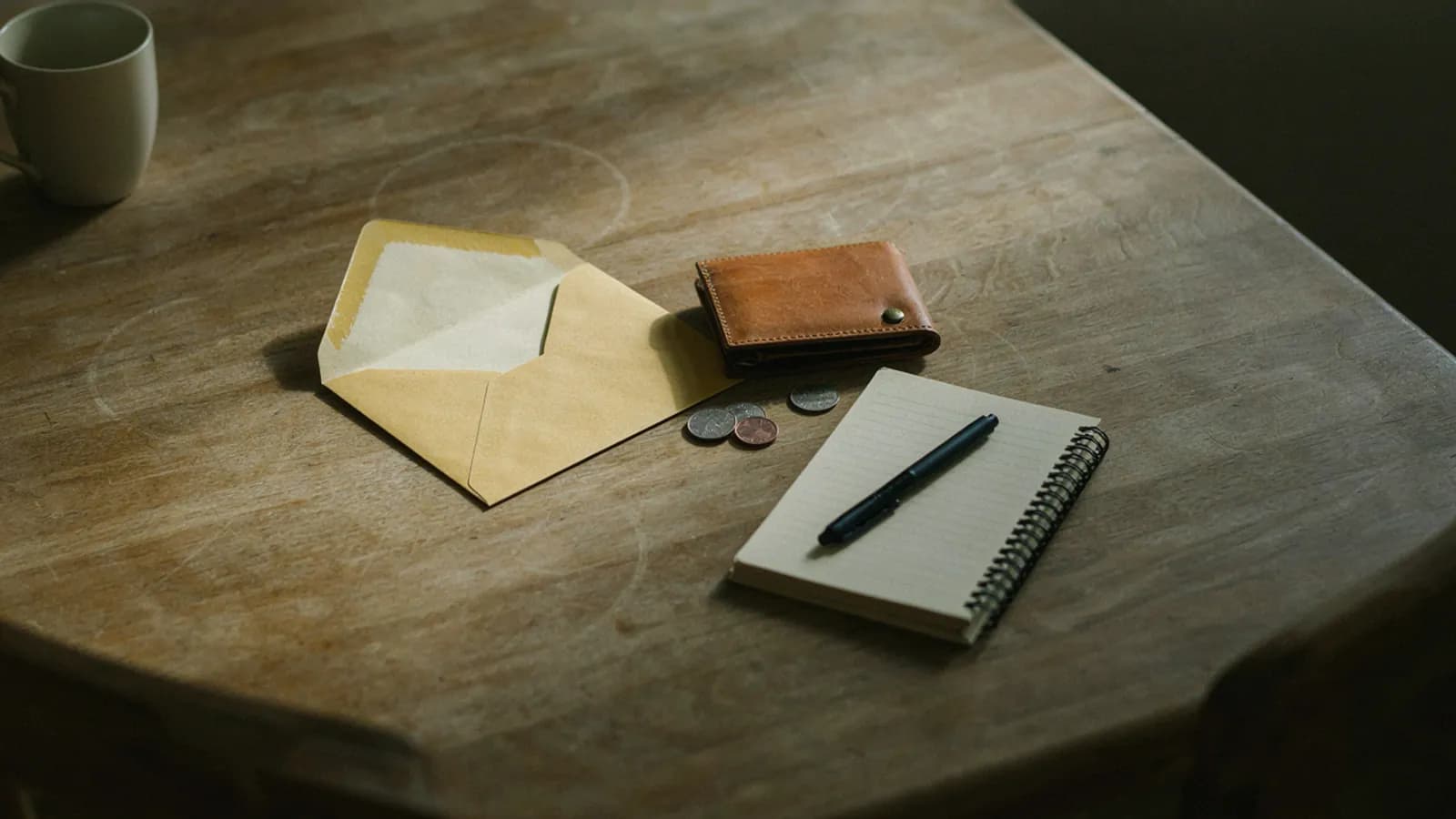 A pay envelope, a leather wallet, a few coins and a notebook with a pen on a wooden table; AI-generated illustration