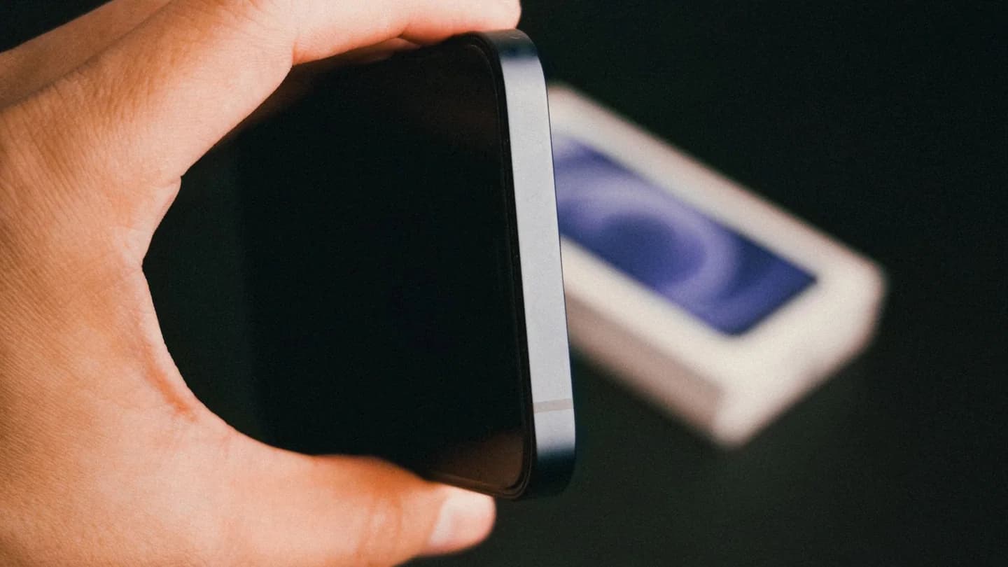 A hand holding a dark smartphone by its edge above a blurred retail box; illustrative photo, not the foldable iPhone Duo