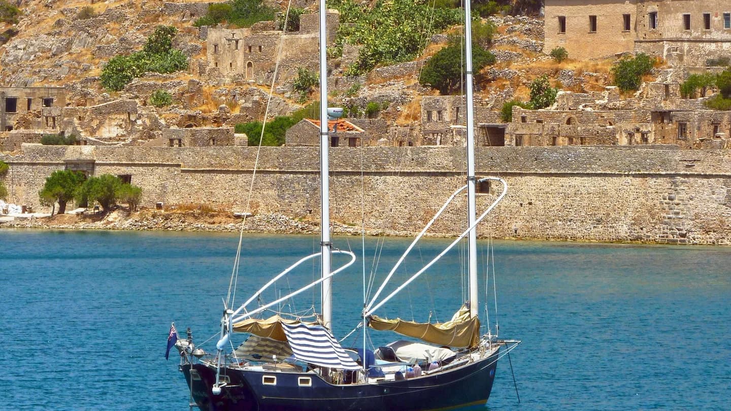 A two-masted sailing boat anchored in blue water below a rocky Greek island with stone ruins; illustrative photo, not a scene from The Odyssey