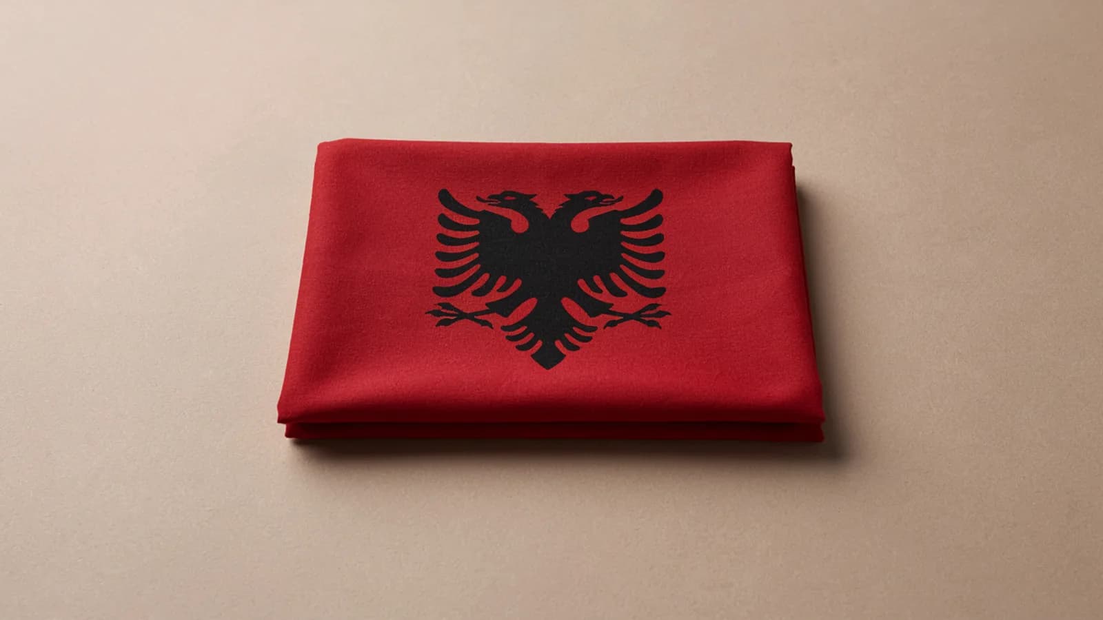 A folded red flag of Albania with the black double-headed eagle on a plain beige background; AI-generated image