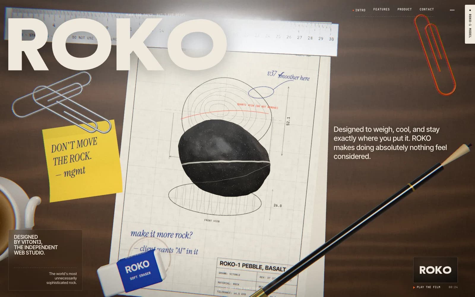 The ROKO home screen: a top-down walnut desk with a technical drawing of the pebble under a real-time 3D basalt stone with a white vein, paperclips, a ruler, a pencil, an eraser and a yellow sticky note reading “Don’t move the rock. — mgmt”, under the huge ROKO wordmark.