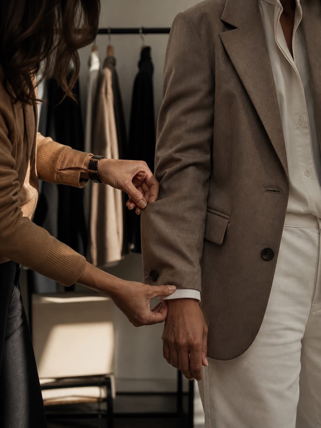 A fitting: a stylist adjusts the sleeve of a brown jacket on a client, with a clothing rail behind them
