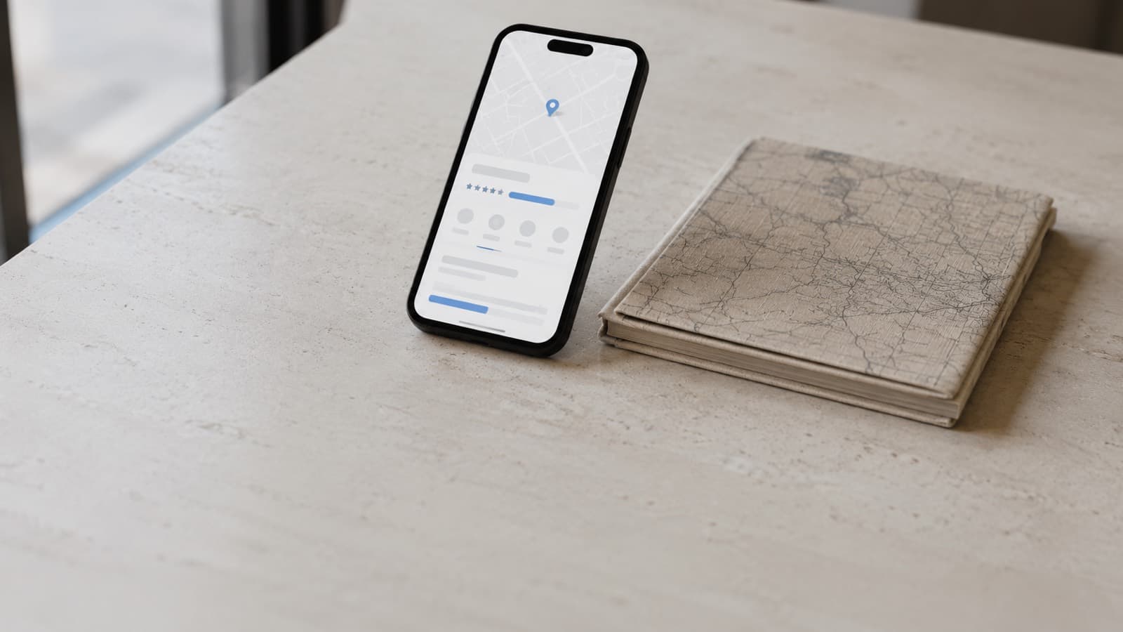 A phone standing on a pale counter showing a map with a pin and a business listing with star ratings, beside a folded paper road map