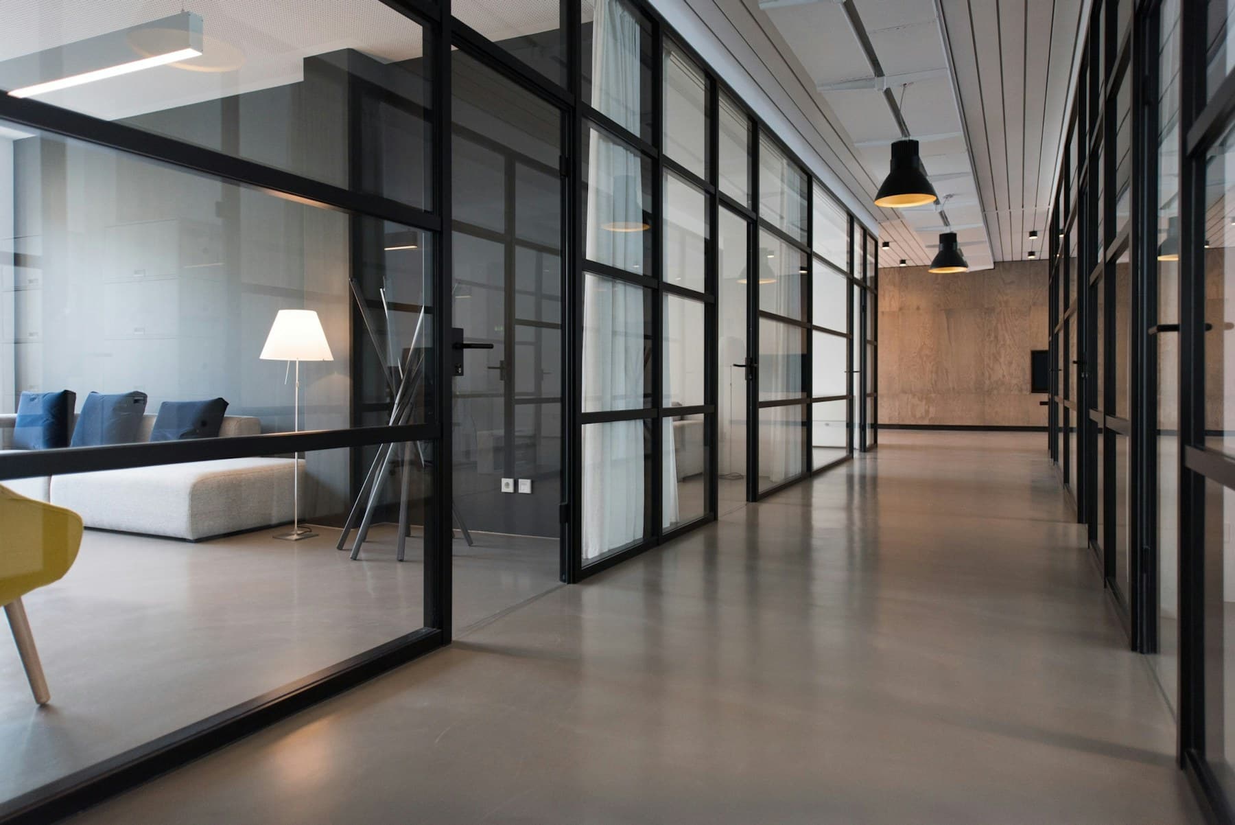 An empty office corridor with black-framed glass partitions, pendant lamps overhead and a lounge visible through the glass