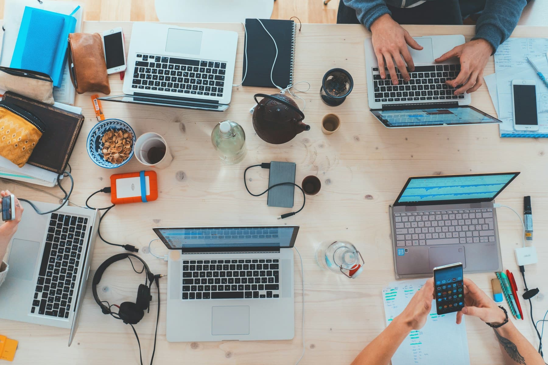Overhead view of a long pale wooden table with four open laptops worked on by several people, with phones, notebooks, headphones, external drives, a teapot and cups between them
