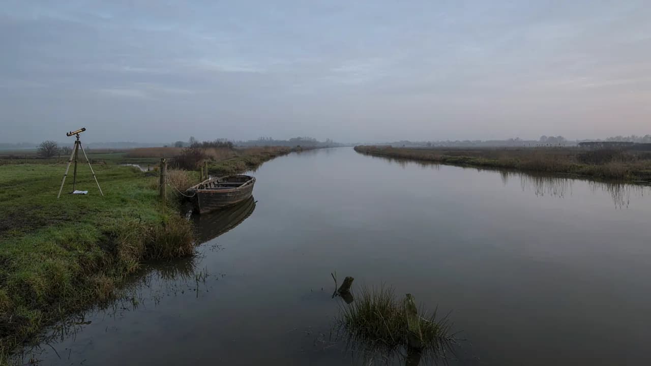 A long straight canal running to the horizon across misty fenland at dawn, with a moored rowing boat and a telescope on a tripod