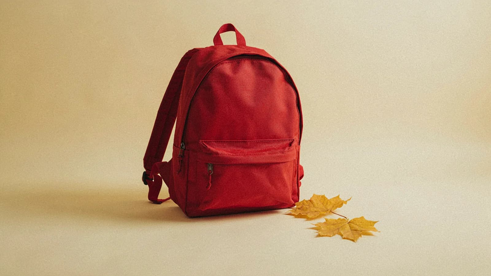 A red school backpack with two yellow maple leaves beside it on a plain warm cream background; AI-generated image