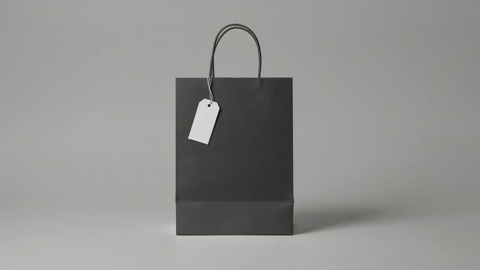 A black paper shopping bag with a blank white tag on its handle, on a plain graphite grey background; AI-generated image