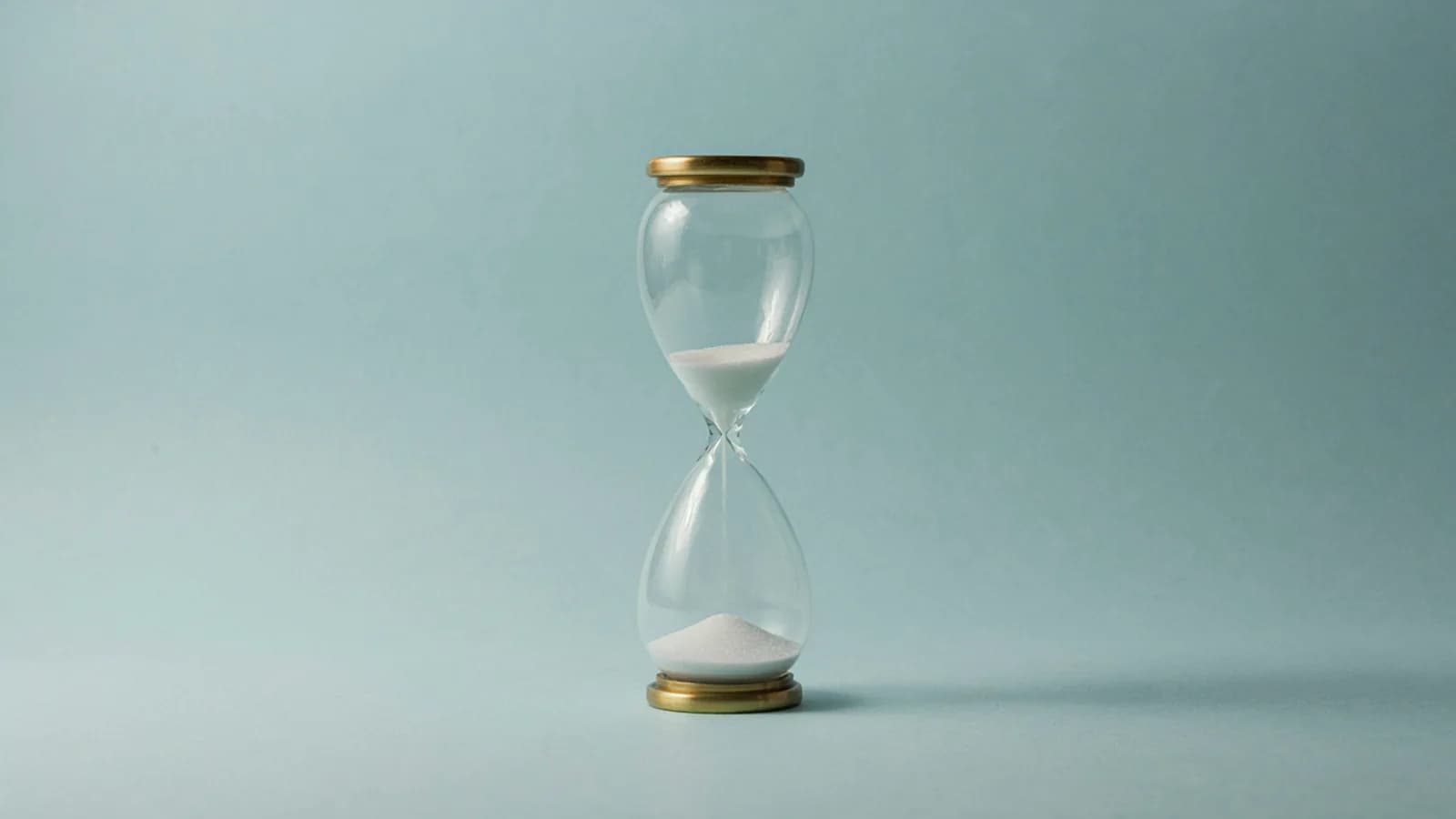 A glass hourglass with brass ends, part of its white sand already fallen, on a plain pale blue-green background; AI-generated image